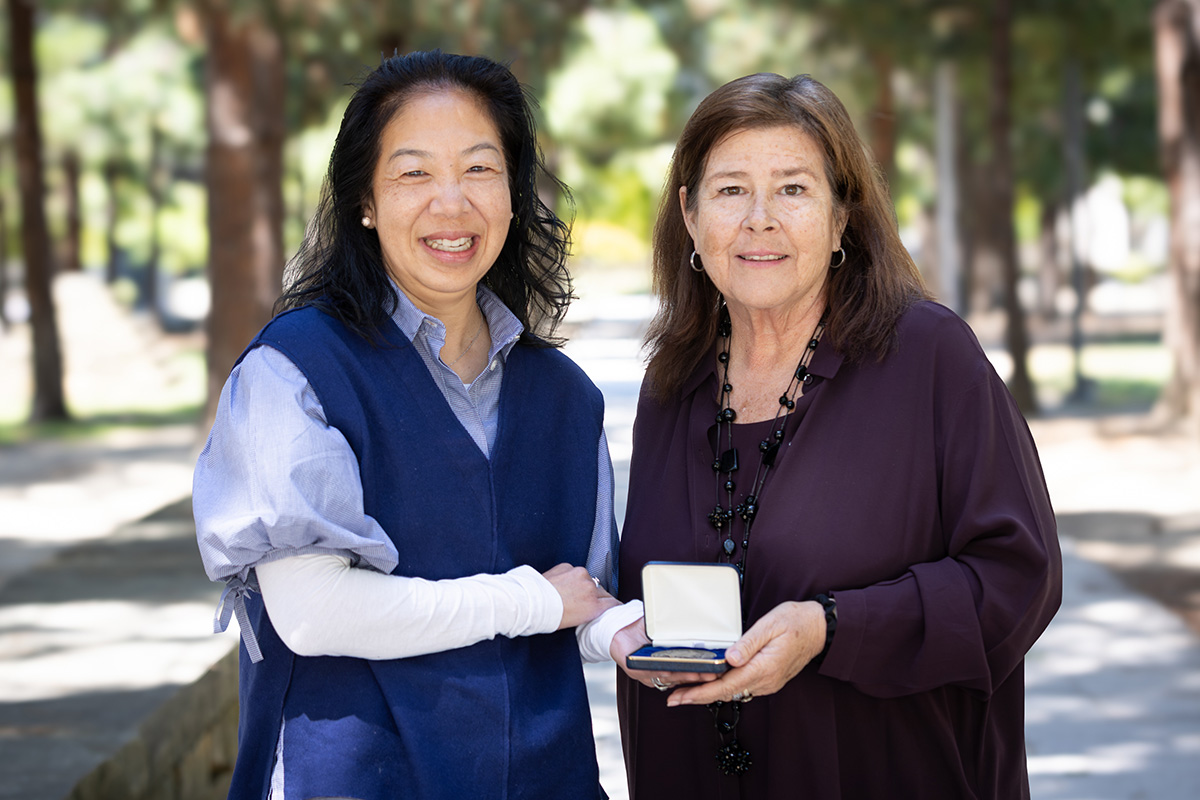 Sabrina Wong stands next to Carol Dawson-Rose holding a small open box displaying a medal. 