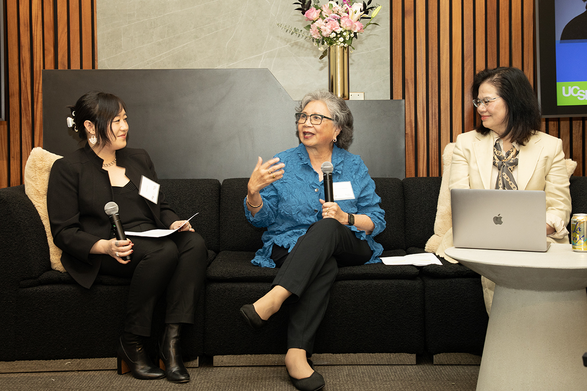 Jennie Chin Hansen, seated on a black couch, speaks into a microphone and gestures while Jarmin Yeh and Jian Zhang, seated on either side of her, look at her and smile. 