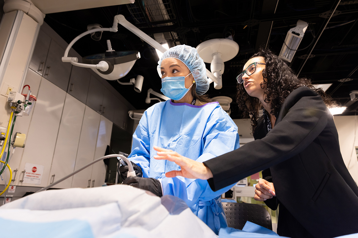 A student in scrubs, hair covering, surgical mask, and gloves, holds a scope over a draped form as a woman in suit jacket leans over to guide her hand. Both are looking up and to the left, like they are watching a screen.