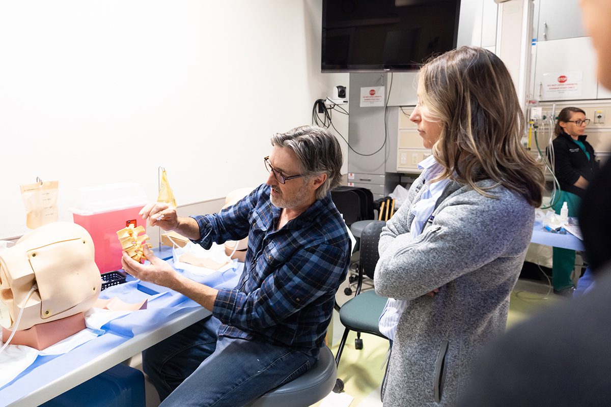 A standing woman in a gray jacket watches as a seated man guides a needle toward a model of a segment of spine. 