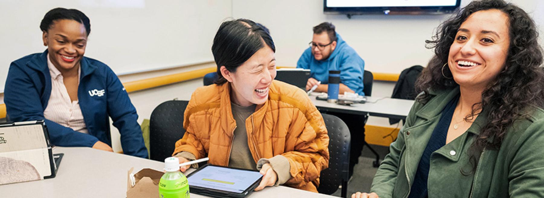 Three students sit in class, laughing and talking, with one student sitting in the background.