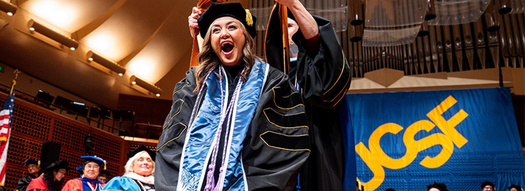 A student stands on the stage at commencement and is hooded by a faculty member who is standing behind them. A banner with the letters "UCSF" is behind them.