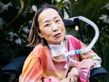 Asian American disability rights activist Alice Wong sits in her wheelchair with a tracheostomy at her neck connected to a ventilator. She’s wearing a pink plaid shirt and a magenta lip color. She is smiling and behind her is vibrant green foliage. Photo credit: Allison Busch Photography.)