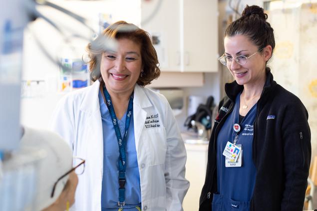 Two nurses smiling down at a patient in a bed. The nurse on the left is wearing a white coat and a UCSF Health lanyard. 