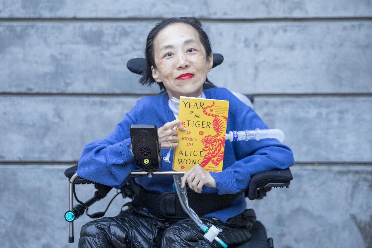 Photo of Alice Wong in a power chair. She is wearing a blue sweater. She is holding a copy of her memoir, Year of the Tiger. In the background is a gray cement wall. 