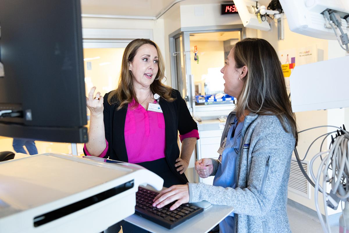 Two women stand together at a computer in a hospital suite. One gestures at the screen while the other looks on. 