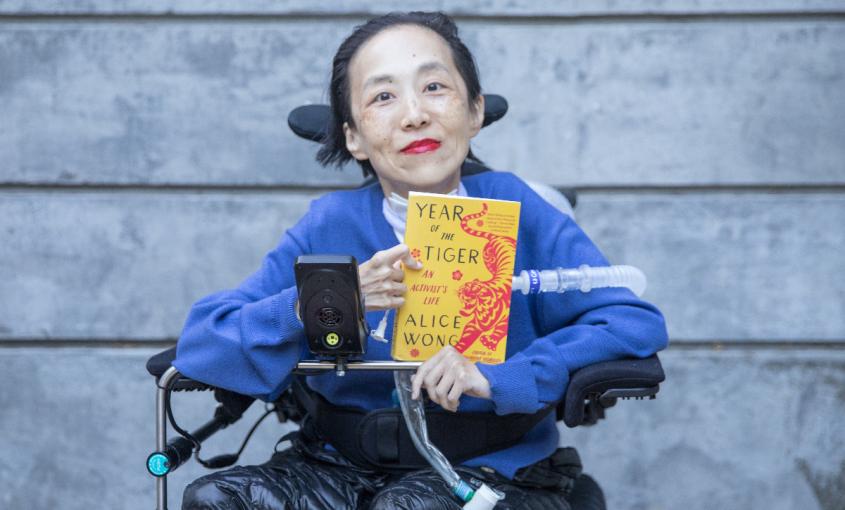 Photo of Alice Wong in a power chair. She is wearing a blue sweater. She is holding a copy of her memoir, Year of the Tiger. In the background is a gray cement wall. 