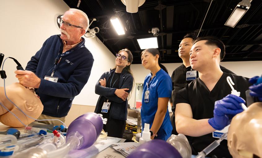 Four students watch as a professor demonstrates a procedure in a simulation lab.