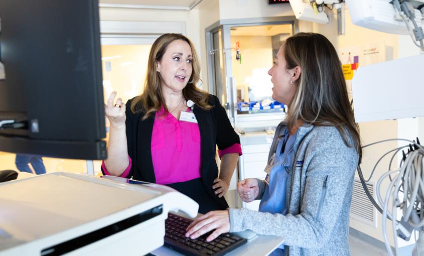 Two women stand together at a computer in a hospital suite. One gestures at the screen while the other looks on. 