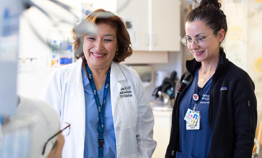 Two nurses smiling down at a patient in a bed. The nurse on the left is wearing a white coat and a UCSF Health lanyard. 