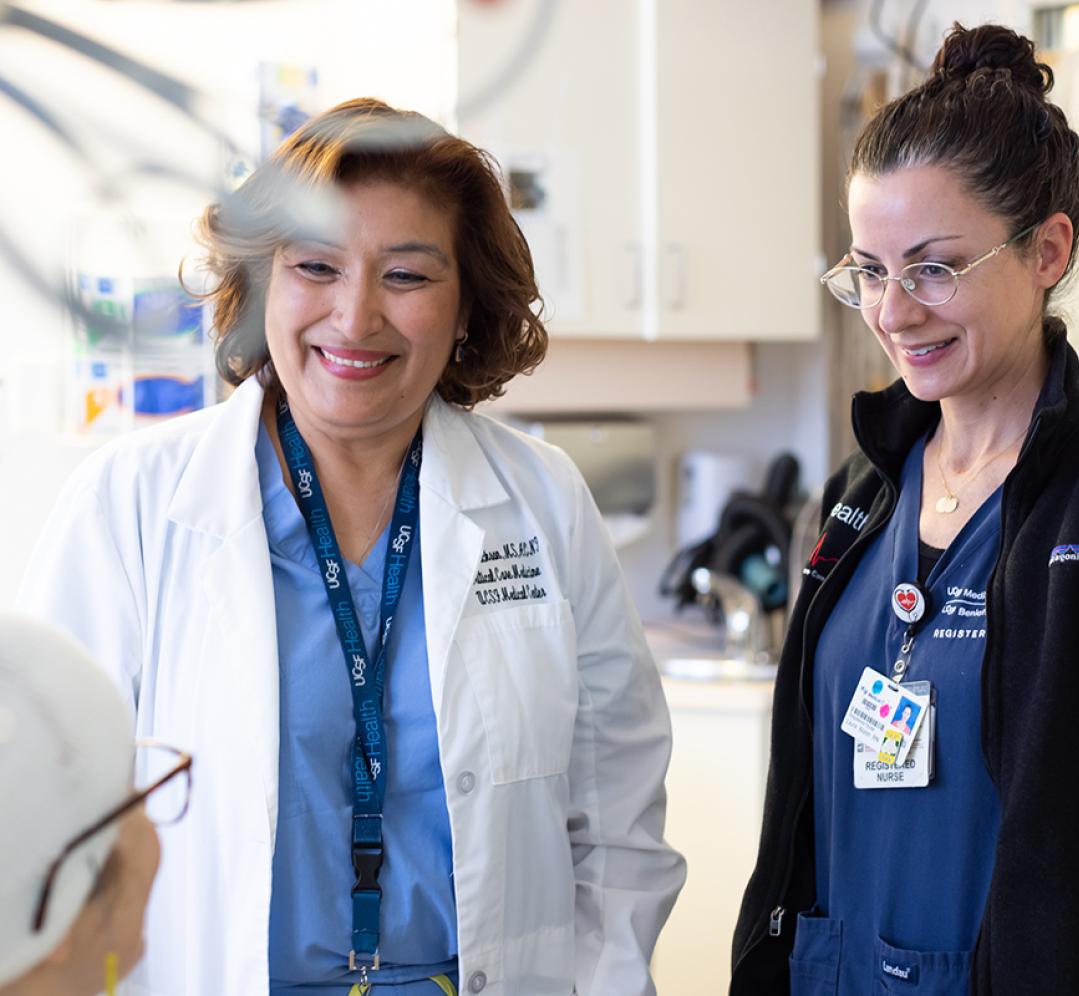 Two nurses smiling down at a patient in a bed. The nurse on the left is wearing a white coat and a UCSF Health lanyard. 